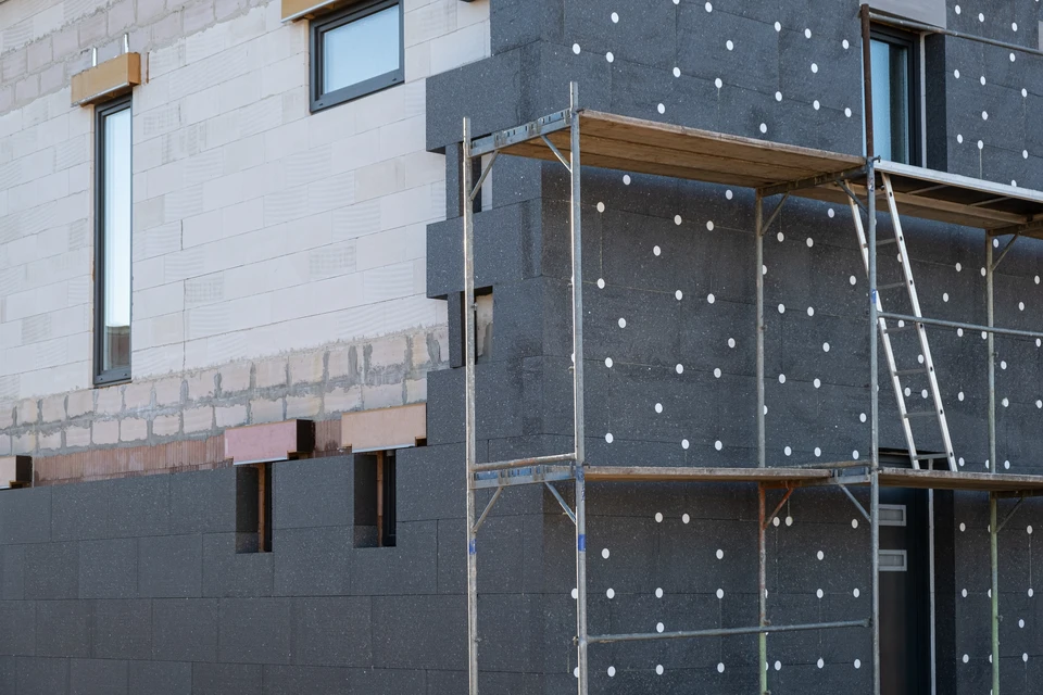 Ouvrier qualifié en tenue de travail appliquant des panneaux d'isolant thermique blanc sur la façade d'une maison résidentielle, avec les rails de départ métalliques visibles et pré-installés. La scène doit montrer la phase intermédiaire de travail d'ITE : les panneaux d'isolant en cours de pose, fixation en cours, avant la finition crépi/peinture. L'ouvrier utilise les équipements professionnels appropriés (niveau, outils de fixation). La maison en arrière-plan doit être clairement une habitation résidentielle individuelle typique de la région Alsace.