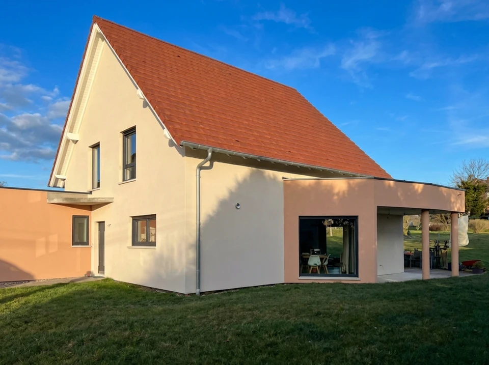 Photographie réaliste montrant la façade avant et après d'une maison individuelle ayant subi un ravalement complet. L'image doit présenter un ouvrier ou une équipe appliquant un enduit ou une peinture sur la façade extérieure d'une maison résidentielle en Alsace, avec des outils professionnels (taloche, brosse, applicateur). La façade partiellement rénovée doit montrer clairement la différence entre la partie restaurée (propre, lisse, peinte, valorisée) et l'ancienne partie usée, démontrant ainsi la transformation et la valorisation du bien immobilier. L'image doit capturer le travail professionnel d'un ravalement complet incluant le rebouchage, le nettoyage et la finition extérieure.